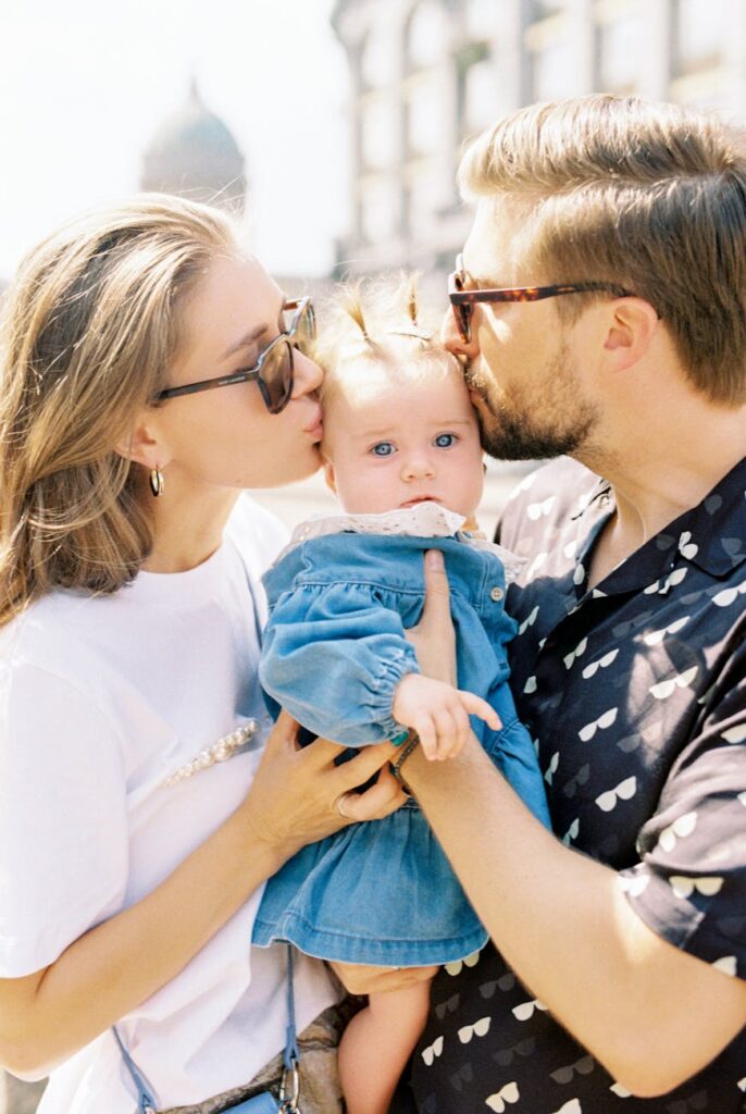 A joyful family portrait with parents kissing their baby outdoors on a sunny day.